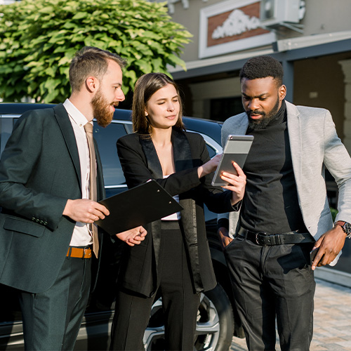 Two men, African and Caucasian, and one Caucasian woman stand in front of black car on a parking place on yard of dealer. Woman showing something on the digital tablet.