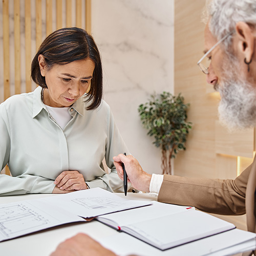 bearded realtor with tattoo pointing with pen at blueprint near woman buying real estate property