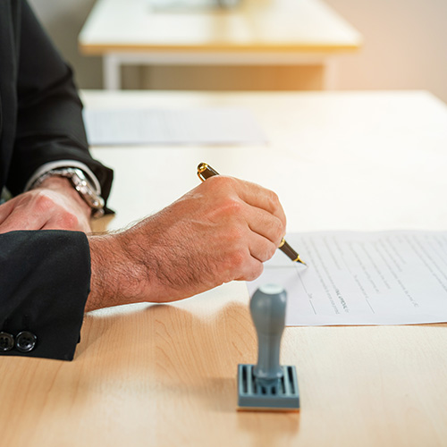 Close-up of a person's hand sign with approved on certificate document at desk.