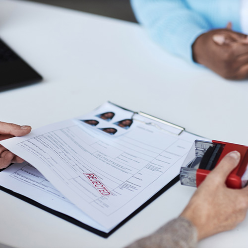 Hands of female manager of visa application center putting rejected stamp on filled form of African American applicant sitting in front of her